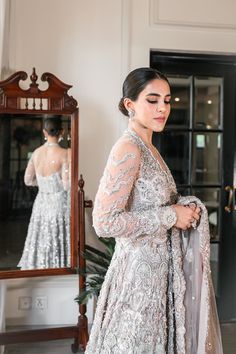 Pakistani Bridal Walima Dress in Gown Lehenga Style has an alluring silver-grey shade and it is gracefully emblazoned with intricate designs and fine details of embellishments. Perfect stitching and premium quality fabric make this Pakistani Bridal Dress an epitome of beauty. Pakistani Bridal Gown: Pakistani Bridal Gown is adorned with hand-crafted details of beaming silverwork, zardosi, pearls, and sequins. This Pakistani Bridal Dress is fully emblazoned with shimmering ornaments and motifs, ma Silver Dress With Zari Work For Eid, Hand Embellished Silver Dress For Eid, Silver Eid Dress With Zari Work, Silver Hand Embellished Dress For Eid, Silver Hand Embellished Sharara For Reception, Festive Silver Dress With Resham Embroidery, Festive Silver Dress With Mirror Work, Festive Silver Hand Embellished Dress, Elegant Silver Choli For Eid