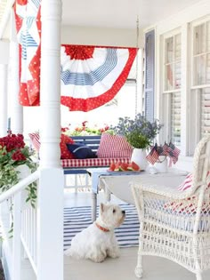 a white dog sitting on the porch next to a table and chairs with red, white and blue decorations