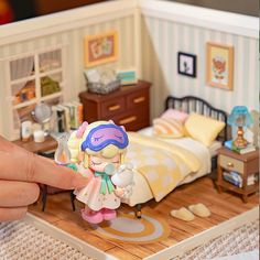 a hand is holding a doll in front of a miniature bedroom