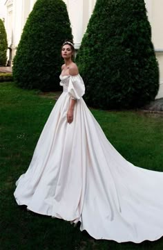 a woman in a white wedding dress standing on the grass near some bushes and trees