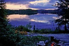 a campfire in front of a lake with trees on the shore and clouds in the sky