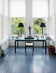 a table with two lamps and some pictures on it in front of three windows that look out onto the woods