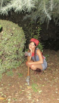 a woman kneeling down next to a bush with a shovel in her hand and wearing a red hat