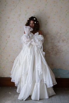 Evocative of an angelic 1960s ceremonial gown, this resplendent white confection radiates the unbridled joy and light of coming-of-age. Layers of billowing organdy drift like clouds over the pure silhouette, while the long sleeves, adorned with ties and lace trim, mirror the cinched details of the skirt. The off-the-sh