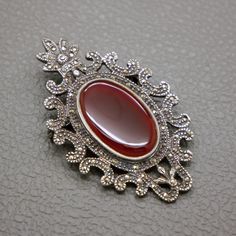 This exquisite vintage brooch is a true statement piece, featuring a striking brown-red carnelian gemstone set in sterling silver. Crafted circa 1950s -1970s, this brooch showcases a timeless Edwardian style adorned with sparkling marcasites. The intricate silver work beautifully complements the smooth, polished carnelian, making it a perfect addition to any vintage jewelry collection. Whether worn on a jacket lapel, dress, or scarf, this large silver brooch adds a touch of elegance and sophisti Vintage Gemstone Brooches For Formal Occasions, Vintage Gemstone Brooches, Ornate Gemstone Brooches For Collectors, Vintage Silver Brooch With Gemstone, Vintage Silver Gemstone Brooch, Antique Pendant Brooches With Gemstone, Elegant Red Brooches Collectible, Ornate Red Jewelry Brooch, Vintage Carnelian Jewelry For Weddings