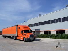 an orange semi truck is parked in front of a large building with windows on the side