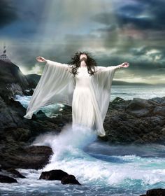 a woman in white dress standing on rocks near the ocean with her arms spread out