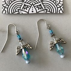 two pairs of earrings with blue beads and angel wings on them, sitting next to a piece of artwork