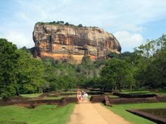 people walking down a dirt path in front of a large rock formation