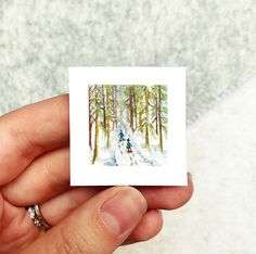 a hand holding a small card with a painting of two people walking in the woods