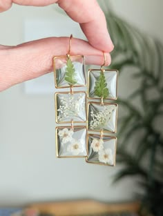 a person is holding four square earrings with flowers and leaves in them on a table