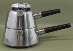 a stainless steel tea kettle with two black handles and one silver handle, on a green surface