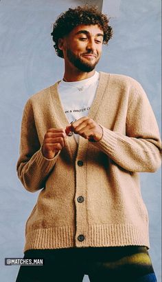 a man standing in front of a blue background wearing a tan cardigan and black pants