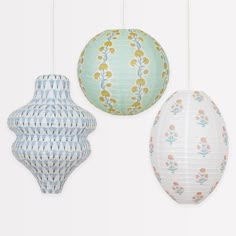 three paper lanterns hanging from strings in various patterns and colors on a white wall behind them is an ornament