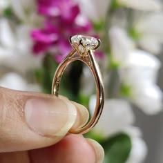 a person holding a gold ring with two diamonds on it and flowers in the background