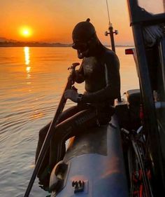 a man sitting on the back of a boat while holding onto a fishing pole with the sun setting in the background