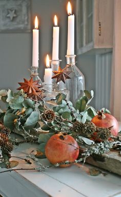 an arrangement of candles, apples and greenery on a table in front of a window