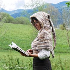 a woman wearing a knitted hooded shawl reading a book in a grassy field