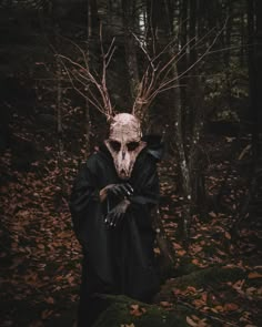 a man in a black robe and mask with horns standing in the woods, holding his hands on his face