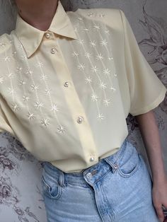 This vintage blouse is made from a delicate pale yellow fabric, featuring short sleeves and adorned with intricate embroidery. The feminine design is highlighted by elegant pearl buttons that add a touch of sophistication. Ideal for both casual and dressy occasions, this shirt embodies the timeless charm of vintage fashion with its detailed craftsmanship and classic silhouette. - Good vintage condition  - TAG SIZE: 44 - Fabric information: 100% polyester  Estimated to fit XS-S-M-L based on your Formal Embroidered Short Sleeve Blouse, Formal Short Sleeve Embroidered Blouse, Elegant Short Sleeve Blouse With Buttons, Fitted Summer Blouse With Pearl Embroidery, Elegant Yellow Summer Shirt, Fitted Pearl Embroidered Blouse For Summer, Classic Tops With Pearl Buttons For Spring, Feminine Short Sleeve Shirt With Buttons, Cream Formal Top For Summer