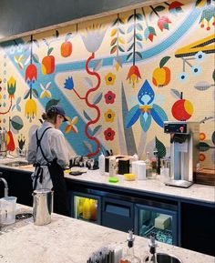 a woman standing in front of a colorful mural behind a kitchen counter with a coffee maker on it