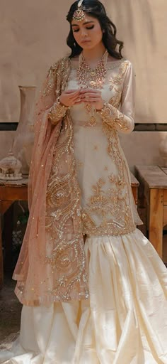 a woman in a white and gold wedding dress is holding her hands together while standing outside