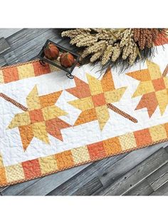 an orange and white quilted table runner with autumn leaves on it, next to pine cones