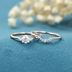 two gold rings with white diamonds on blue cloth next to flower arrangement and pearl necklace