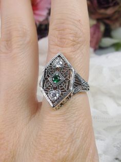 Simulated Green Emerald & White Cubic Zirconia (CZ) Sterling Silver RingCharlotte Design#D231 Resurrected from the Art Deco movement, this gorgeous Art Deco reproduction filigree ring is crafted in sterling silver. This lovely filigree ring is set with a stunning quality 2mm center round-cut simulated green emerald gemstone. Two small white cubic zirconia gemstones also grace the face of the ring, one near each point, each set in the center of a tiny heart. There are 2 filigree accent flowers th Accent Flowers, Art Deco Revival, Art Deco Emerald Ring, Art Deco Movement, Gem Diamonds, Ring Art Deco, Cubic Zirconia Rings, Tiny Heart, Filigree Ring