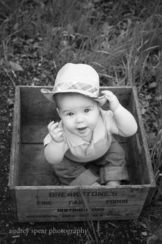 a baby sitting in a wooden box on the ground wearing a hat and looking at the camera