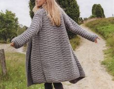 a woman walking down a dirt road wearing a gray coat and black boots with her arms outstretched