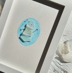 a watercolor painting of a snowman in a frame on a wooden table next to some rocks