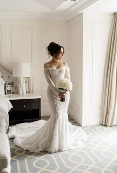 a woman in a wedding dress is standing near a bed and holding a flower bouquet