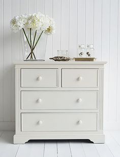 a white dresser with flowers on top and two glasses in the bottom drawer, against a white wall