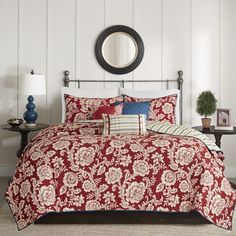 a bed with red and white comforter in a room next to a table, lamp and mirror