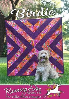 a dog sitting in the grass next to a purple and pink quilted wall hanging