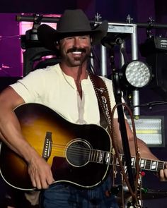 a man wearing a cowboy hat and holding a guitar in front of a microphone on stage