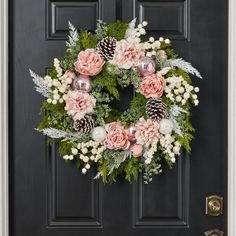 a wreath with pink flowers and pine cones hanging on a black front door decorated with greenery