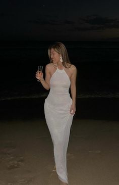 a woman in a white dress on the beach at night holding a glass of wine