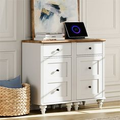 a white dresser with a laptop on top of it next to a basket and painting