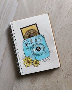 a spiral notebook with an image of a camera and flowers on the cover, sitting on a marble surface