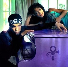 two people leaning on a purple piano in front of a window