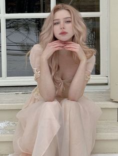 a woman with long blonde hair sitting on a window sill wearing a pink dress