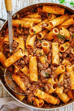 a skillet filled with pasta and meat sauce on top of a wooden table next to a spoon