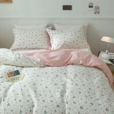 a bed with pink sheets and pillows in a white room next to a lamp on a table