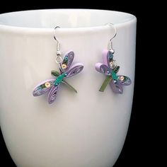 two purple and green dragonfly earrings sitting on top of a white cup in front of a black background