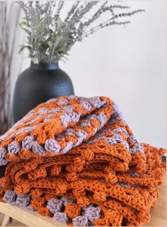 a stack of crocheted blankets sitting on top of a wooden table next to a vase