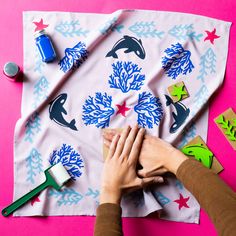 two hands on a piece of fabric with dolphins and corals painted on it, surrounded by other crafting supplies