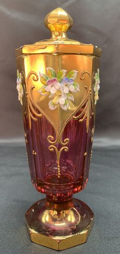 an ornate glass vase with gold trimmings and flowers on the top, sitting on a stand