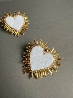 two gold and white heart shaped brooches on a gray surface, one has been made out of beads